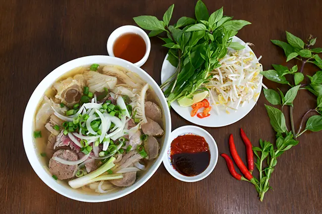 Tô phở bò viên nóng hổi với thịt tái, gân, sách, hành lá, hành tây và rau sống ăn kèm trên bàn gỗ. – Phân loại phở theo vùng miền