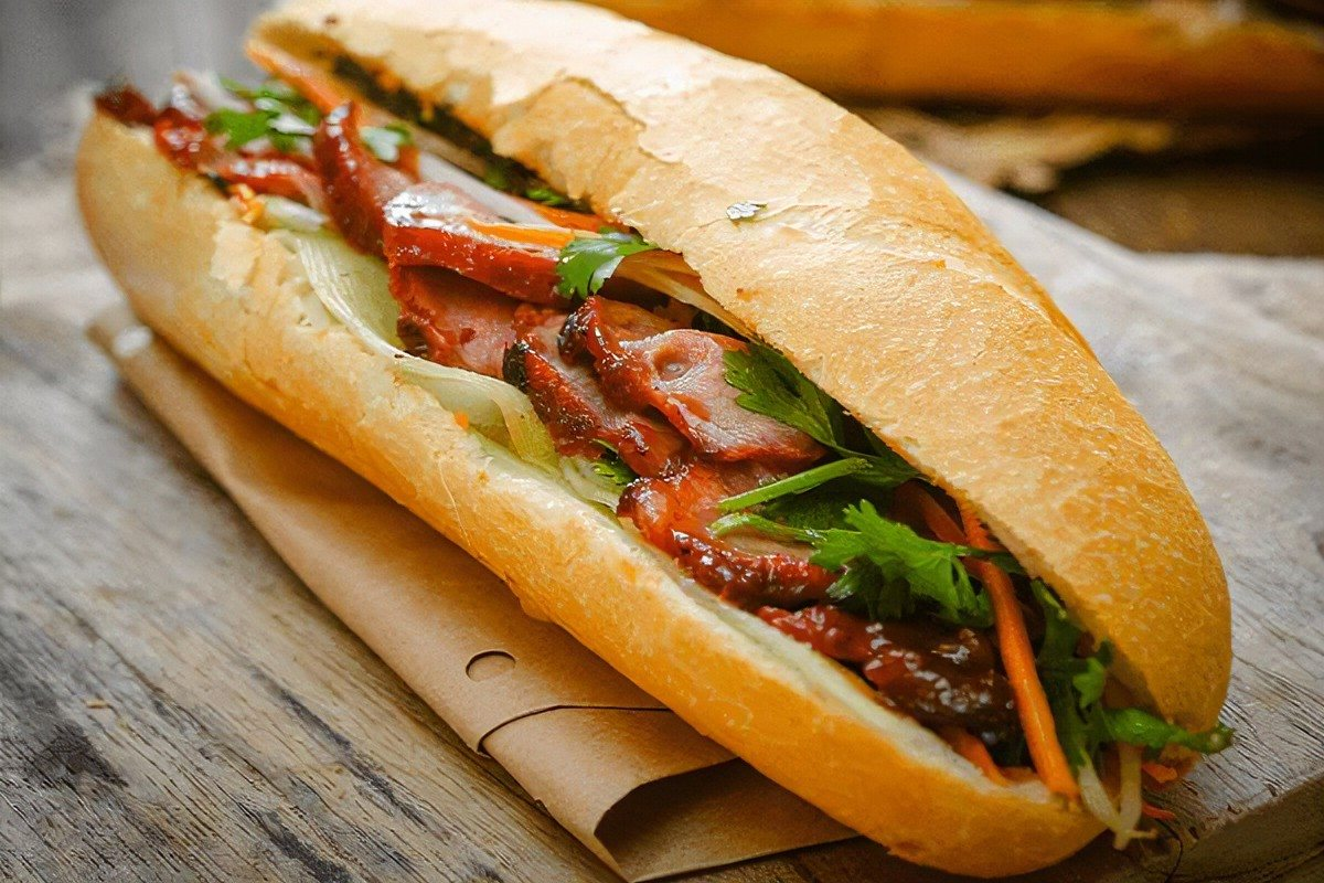 Cận cảnh ổ bánh mì Việt Nam kẹp thịt thái lát, rau mùi, cà rốt sợi và dưa leo đặt trên giấy gói nâu.