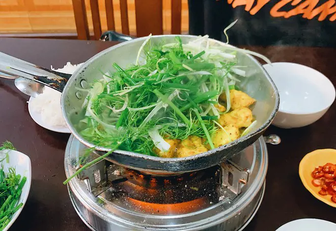 Chảo chả cá Lã Vọng với cá lăng tẩm nghệ, hành hoa và thì là đang sôi trên bếp ga mini tại bàn ăn ở Hà Nội. – Âm thanh xèo xèo trên chiếc chảo gang nhỏ