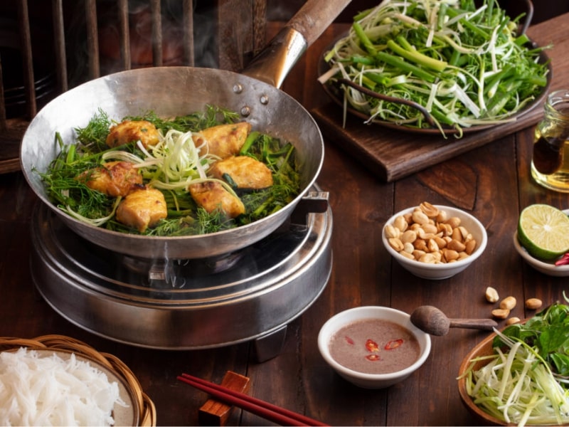 Chảo Chả cá Lã Vọng nóng hổi với cá lăng, thì là và hành lá đang được giữ ấm trên bếp ăn, kèm bún và lạc rang trên bàn ăn nhà hàng.