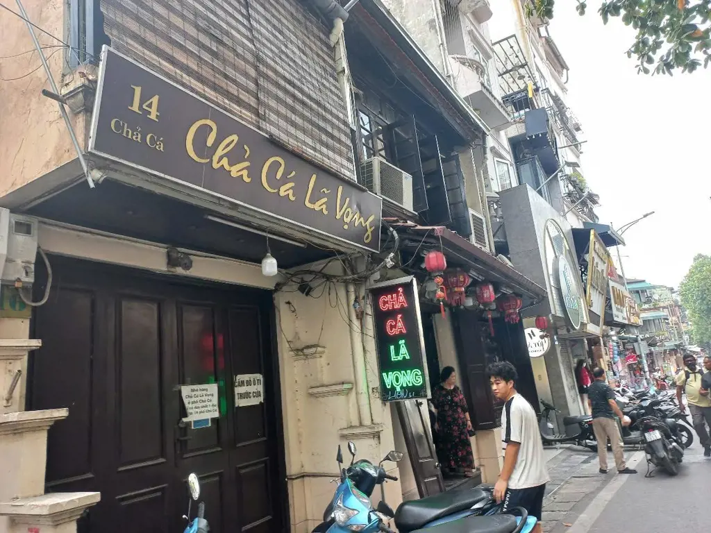 Mặt tiền quán Chả Cá Lã Vọng với biển hiệu số 14 Chả Cá trên một con phố nhỏ ở Hà Nội, phía trước có xe máy và người qua lại.
