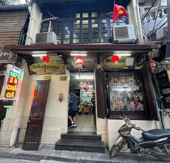 Mặt tiền quán Chả Cá Lã Vọng trong dãy nhà phố ở Hà Nội, treo đèn lồng đỏ, cờ Việt Nam và có xe máy đỗ phía trước.