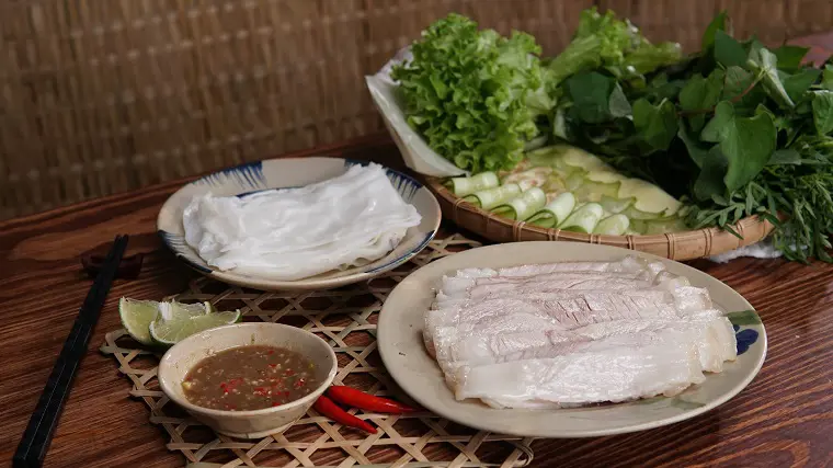 Mẹt bánh tráng thịt heo 2 đầu da kèm đĩa bánh tráng, khay rau sống dưa leo và chén mắm nêm tỏi ớt trên bàn gỗ – Ăn bánh tráng cuốn thịt heo ở đâu?