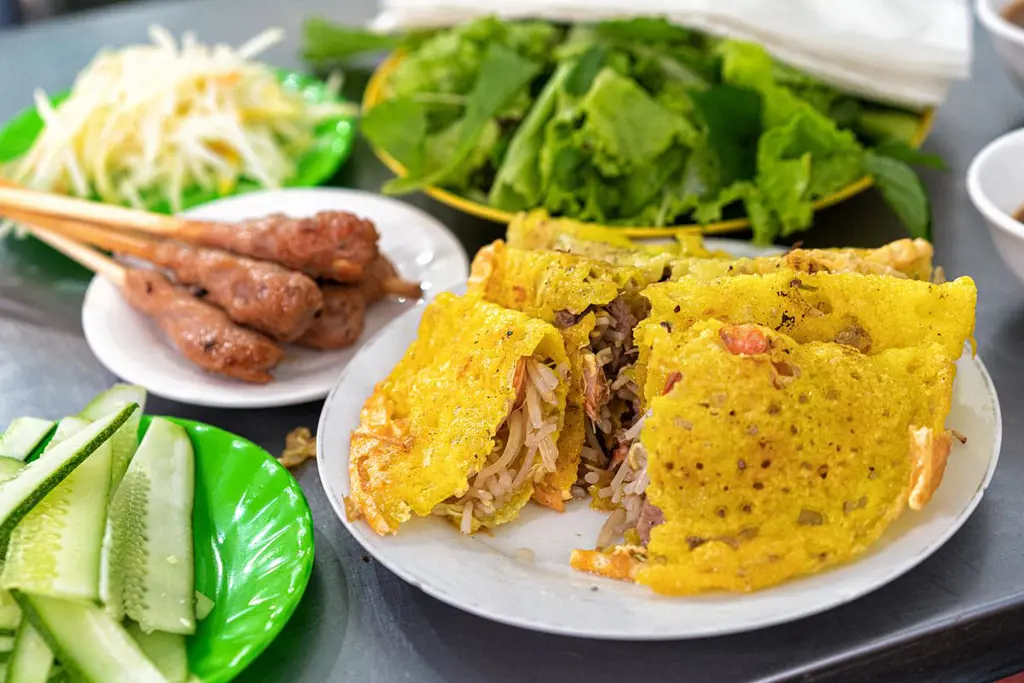 Đĩa bánh xèo vàng giòn cắt đôi kèm nem lụi, dưa leo, đu đủ bào và rau sống trên bàn inox
