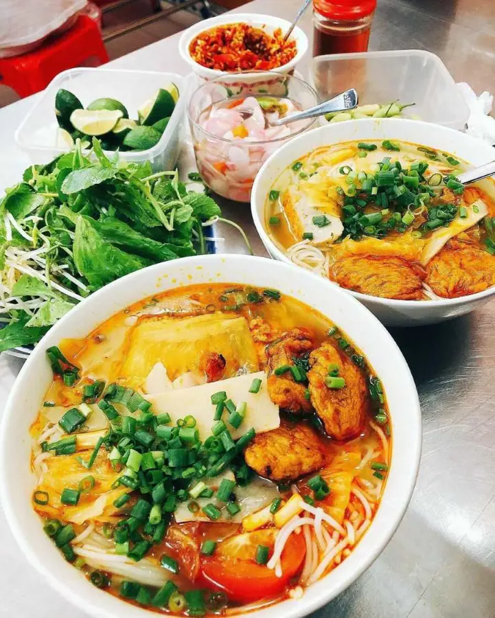 Hai tô bún chả cá Đà Nẵng thơm ngon kèm đĩa rau sống, chanh tươi và các loại gia vị ăn kèm trên bàn inox.