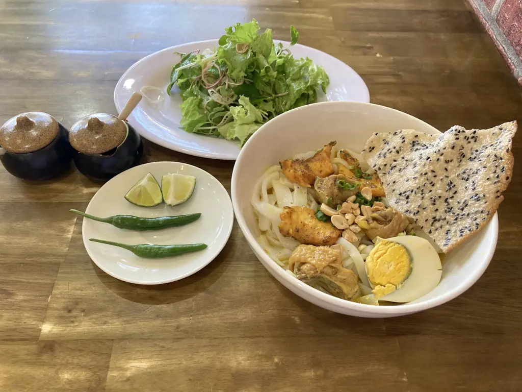 Tô Mì Quảng gà đầy đặn kèm bánh tráng mè nướng, đĩa rau sống, chanh và ớt xanh trên bàn gỗ – 1. Mì Quảng – Linh hồn ẩm thực xứ Quảng