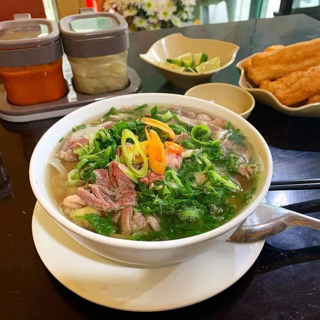 Tô phở bò tái với nước dùng trong, rau thơm, trứng chần, ăn kèm đĩa quẩy và bát ớt tươi thái lát.
