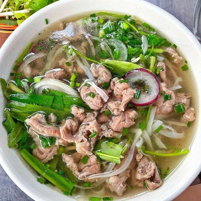 Cận cảnh tô phở bò tái kiểu phở Bắc với thịt bò thái mỏng, nước dùng trong, hành tây, giá đỗ và rau xanh trong bát trắng. – Nước dùng và hương vị thanh mà sâu