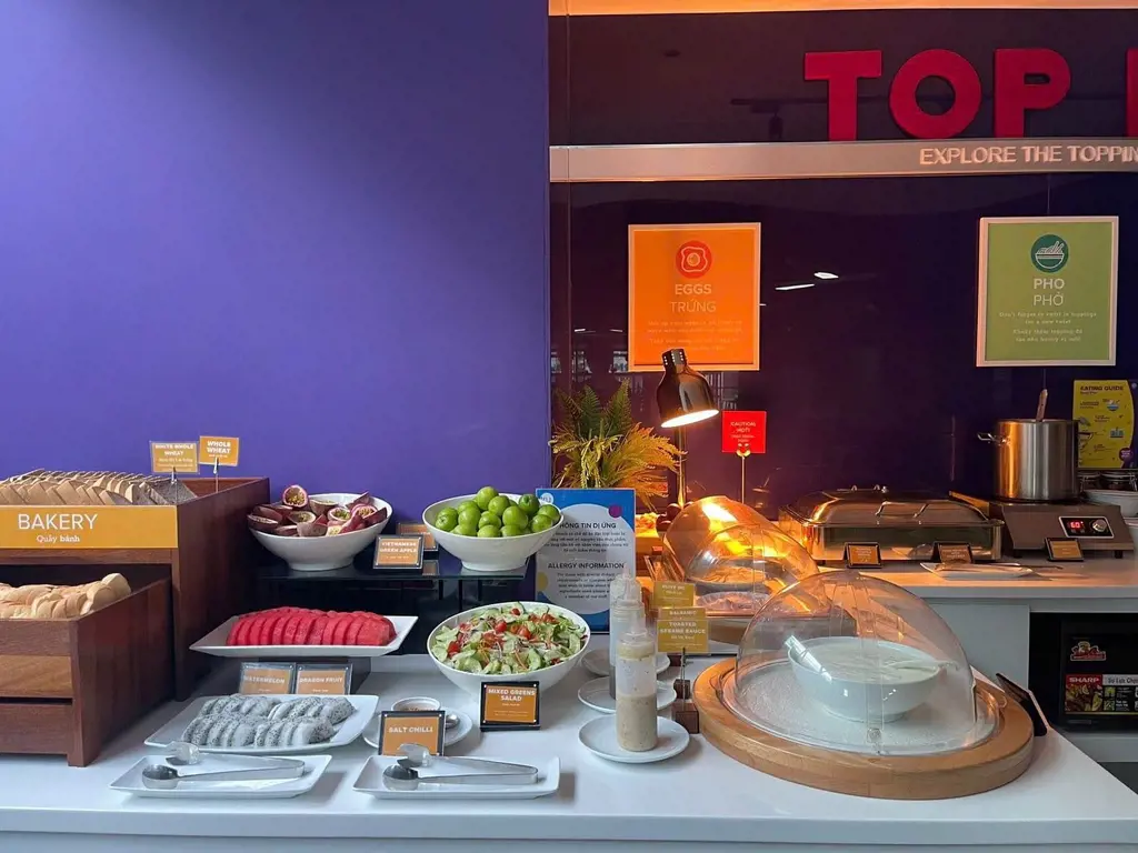 Quầy buffet sáng bày trí trái cây tươi, salad, bánh mì và quầy phở nóng dưới mảng tường tím tại Tru by Hilton Da Nang City Centre