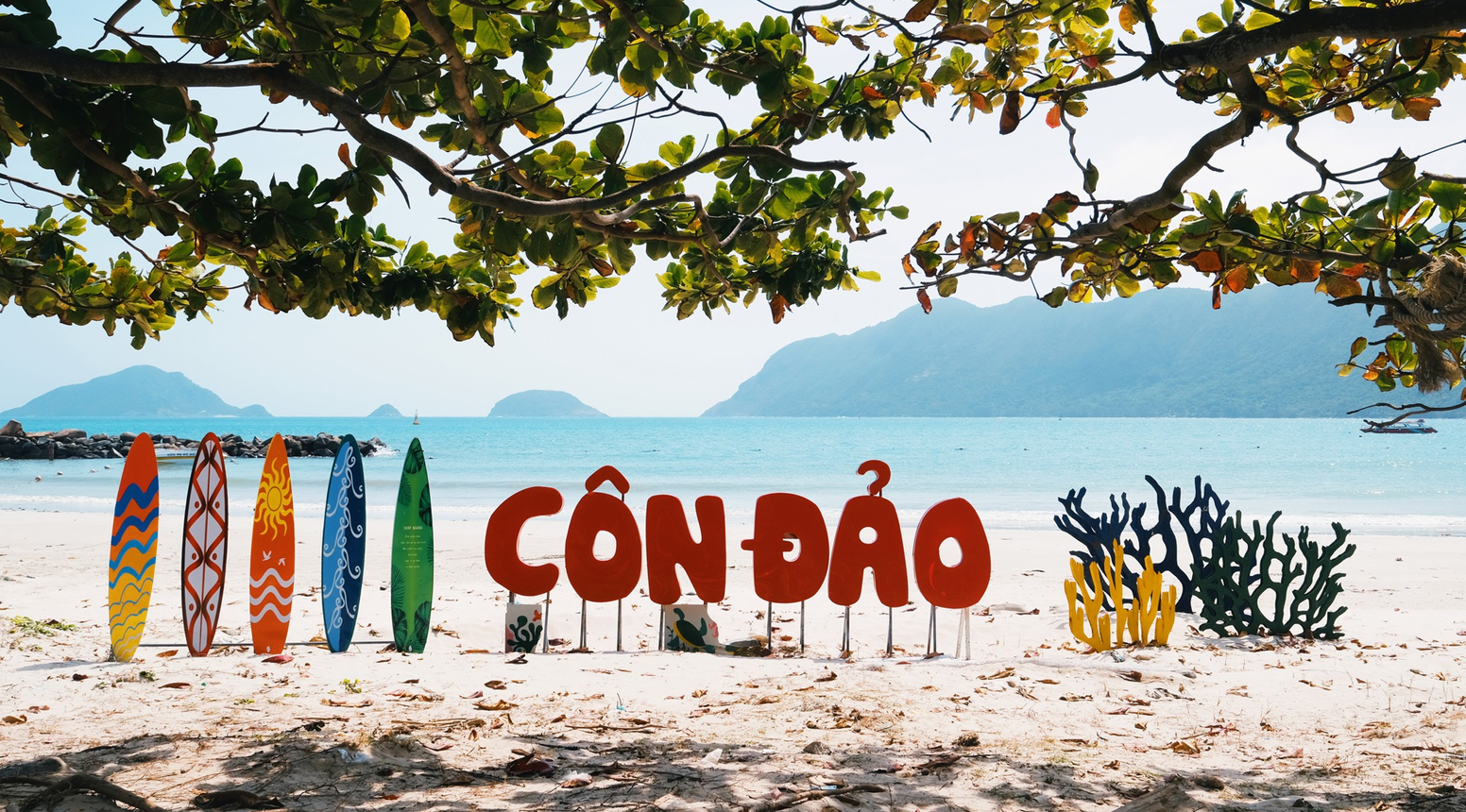 Bảng chữ "CÔN ĐẢO" màu đỏ nổi bật cùng các ván lướt sóng đầy màu sắc trên bãi cát trắng, phía sau là biển xanh và núi non.