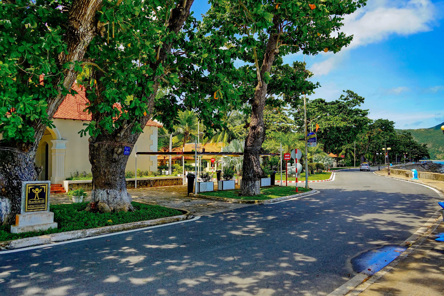 Con đường ven biển rợp bóng cây cạnh khu Dinh Chúa Đảo ở Côn Đảo, bên trái là tòa nhà mái ngói đỏ và bên phải là bờ kè sát mặt nước. – Một ngày như vậy có đáng để thử không?