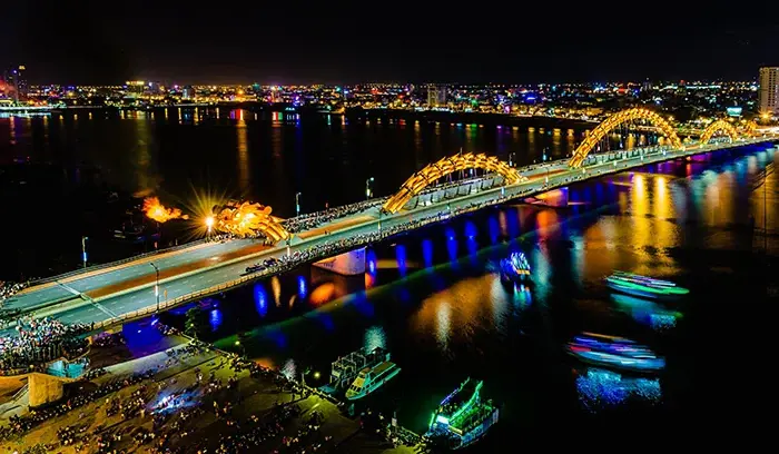 Cầu Rồng Đà Nẵng phun lửa và nước về đêm, với ánh đèn rực rỡ và thành phố lung linh phía sau, nhiều người và thuyền trên sông.