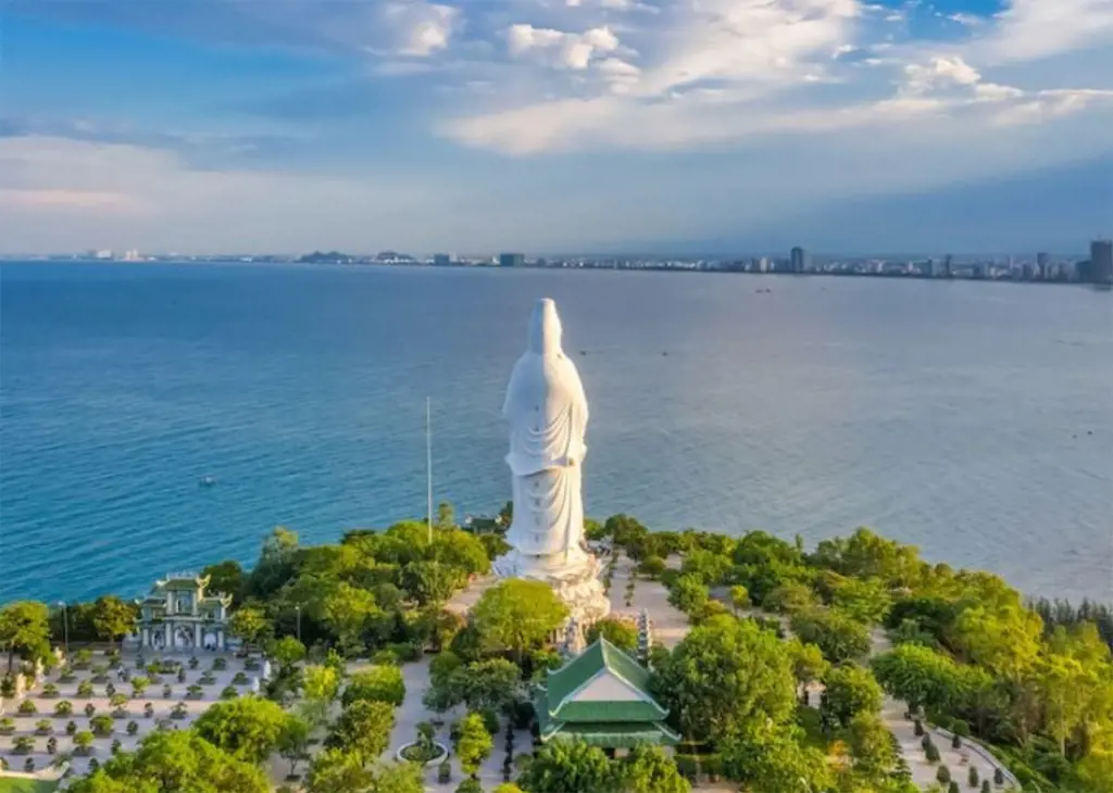 Ảnh chụp từ trên cao tượng Phật Quan Âm màu trắng trong khuôn viên chùa ven biển, phía sau là mặt biển rộng và đường chân trời thành phố, nhiều khả năng là chùa Linh Ứng Sơn Trà ở Đà Nẵng.