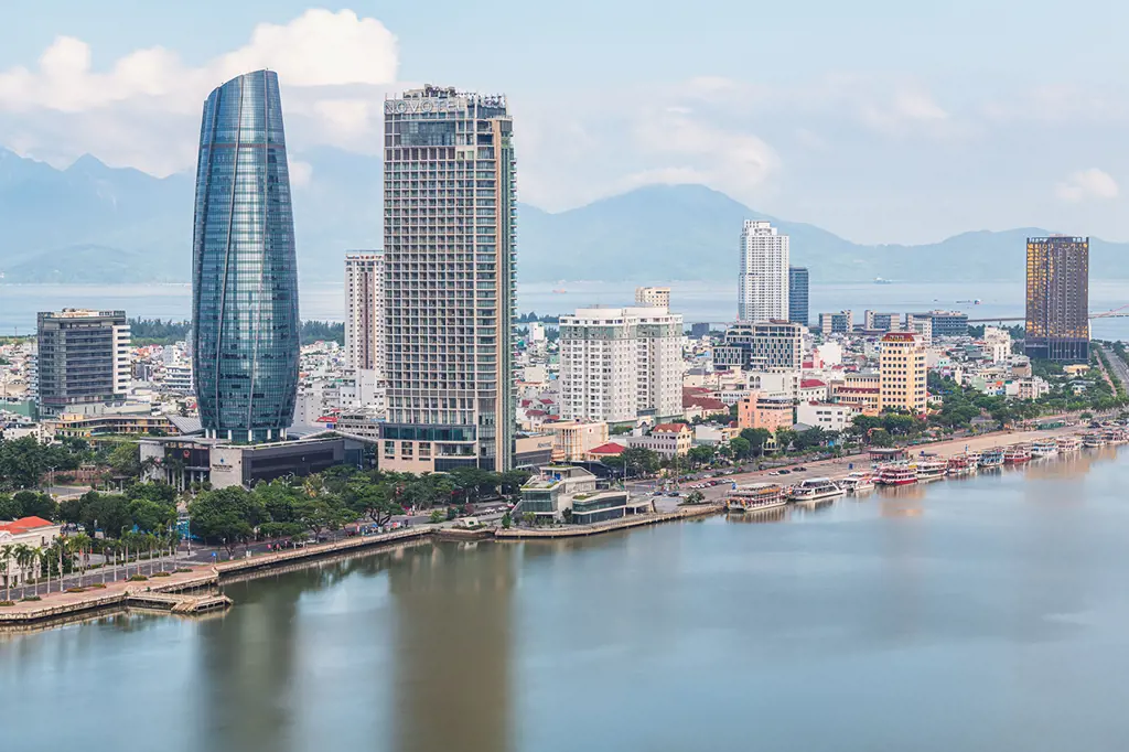 Toàn cảnh các tòa nhà cao tầng ven Sông Hàn ở Đà Nẵng, nổi bật với tháp kính bo tròn, bến thuyền và dãy núi phía xa dưới trời sáng.