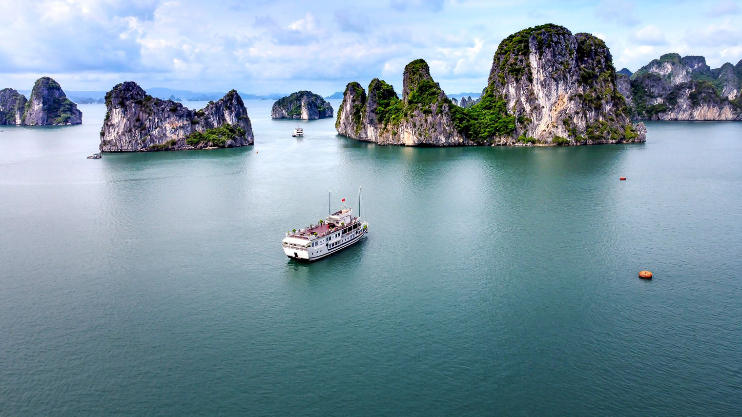 Toàn cảnh Vịnh Hạ Long với nhiều đảo đá vôi xanh tươi, một tàu du lịch lớn màu trắng và nước biển xanh ngọc bích dưới bầu trời trong xanh.