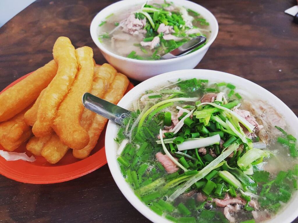 Two bowls of Vietnamese beef pho topped with chopped green onions, served with golden fried dough sticks on a red plate.