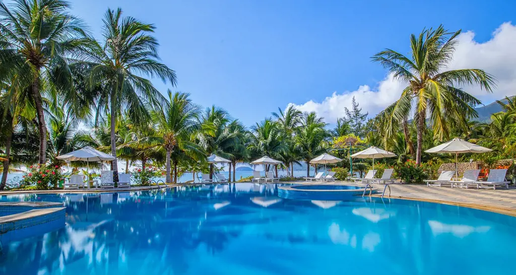 Hồ bơi lớn ngoài trời trong khu Côn Đảo resort ven biển, xung quanh là hàng dừa, ghế tắm nắng và ô che nắng, phía sau nhìn ra biển dưới trời nắng. – 2. Resort có hồ bơi giữa không gian xanh